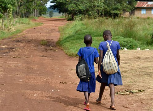 two young girls with blue uniforms and backpacks walking on a dirt road