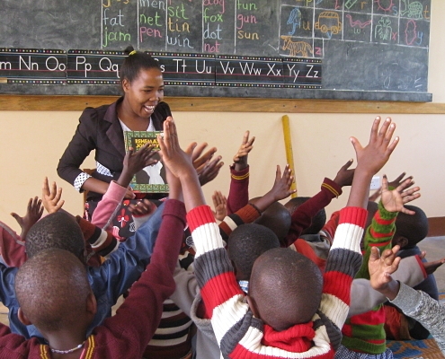 smiling teacher and children with hands raised enthusiastically looking at a book